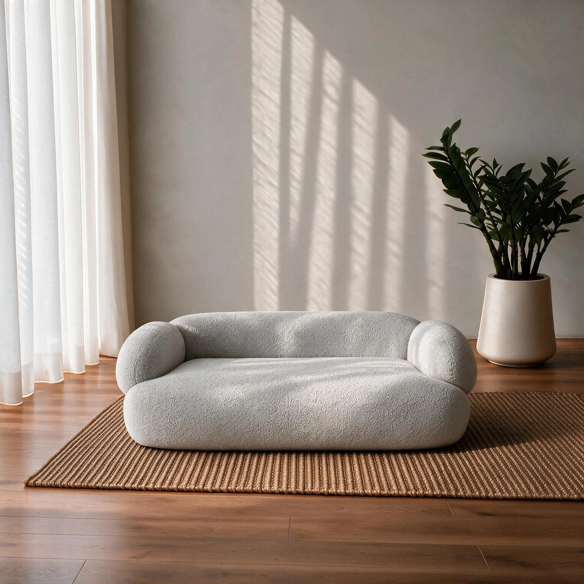 A low, rounded boucle sofa in a light gray color, styled on a woven jute rug in a minimalist room with warm sunlight and shadows.