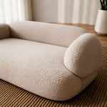 A low, rounded modular sofa or floor cushion in a textured cream bouclé fabric, shown in a softly lit room on a woven brown rug with curtains and a plant in the background.