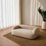 A small rounded sofa or pet bed in a warm beige bouclé fabric, placed on a woven brown rug in a softly lit room with sheer curtains, wood flooring, and a potted plant.