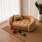 A small tan boucle pet bed with a matching bone-shaped pillow, styled on a woven rug in a bright room with wood flooring, sheer curtains, a potted plant, and a red rope toy. 