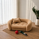A small beige boucle pet bed with a matching cushion, styled on a woven rug in a bright room with curtains, a potted plant, and dog toys on the floor. 