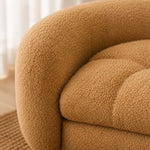 Close-up of a rounded upholstered arm and seat cushion in a warm camel bouclé fabric, with soft natural light and a blurred indoor background. 