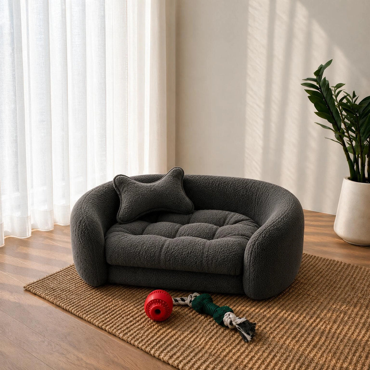 A dark gray plush pet bed with a matching bone-shaped pillow, styled on a woven rug in a bright room with a plant and window curtains. 