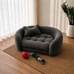 A dark gray plush pet bed shaped like a small sofa with a matching pillow, placed on a woven rug in a sunlit room with a plant and window curtains in the background. 