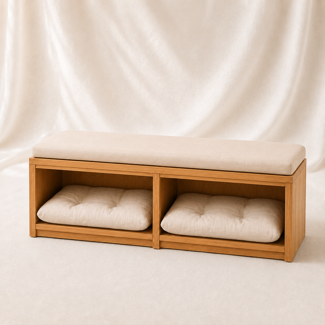 A wooden bench with a thick cream cushion on top and two open storage compartments underneath, each holding a matching cream tufted cushion, set against a draped cream fabric backdrop.