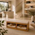 A wooden bench with a cream upholstered seat and two open storage compartments underneath, each holding a small tan dog on a cushion, placed in a bright modern interior with stairs and shelves in the background.