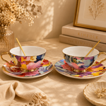 Two floral teacups with saucers on a table with books and decor in the background.