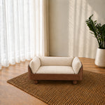 A small wooden pet sofa with cream bouclé cushions sits on a woven jute rug in a sunlit room with sheer curtains and a potted plant.
