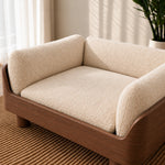 A modern pet sofa chair with a warm brown wood frame and thick, textured cream bouclé cushions, styled in a bright, sunlit room on a woven rug.