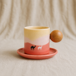 A ceramic coffee cup and saucer set with a warm gradient design of yellow, cream, and coral pink, featuring small camel and deer illustrations. The cup has a round wooden handle, and the background is a soft beige fabric.