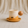 A ceramic coffee mug with a wooden handle shaped like a camel, resting on a matching saucer. The mug features a beige and ochre orange gradient with small camel and llama silhouettes printed on the side, set against a soft beige fabric backdrop.