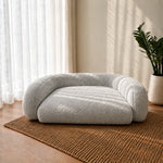 A light gray bouclé pet bed with rounded arms sits on a woven brown rug in a sunlit room with sheer curtains and a potted plant.