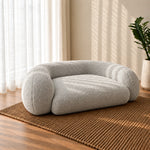 A plush, low-profile round pet bed in light gray bouclé fabric, styled on a brown woven rug in a sunlit room with sheer curtains and a potted plant.