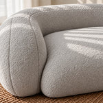 Close-up of a modern curved upholstered chair in a light warm gray bouclé fabric, resting on a woven brown rug with soft sunlight and shadow patterns across the seat.