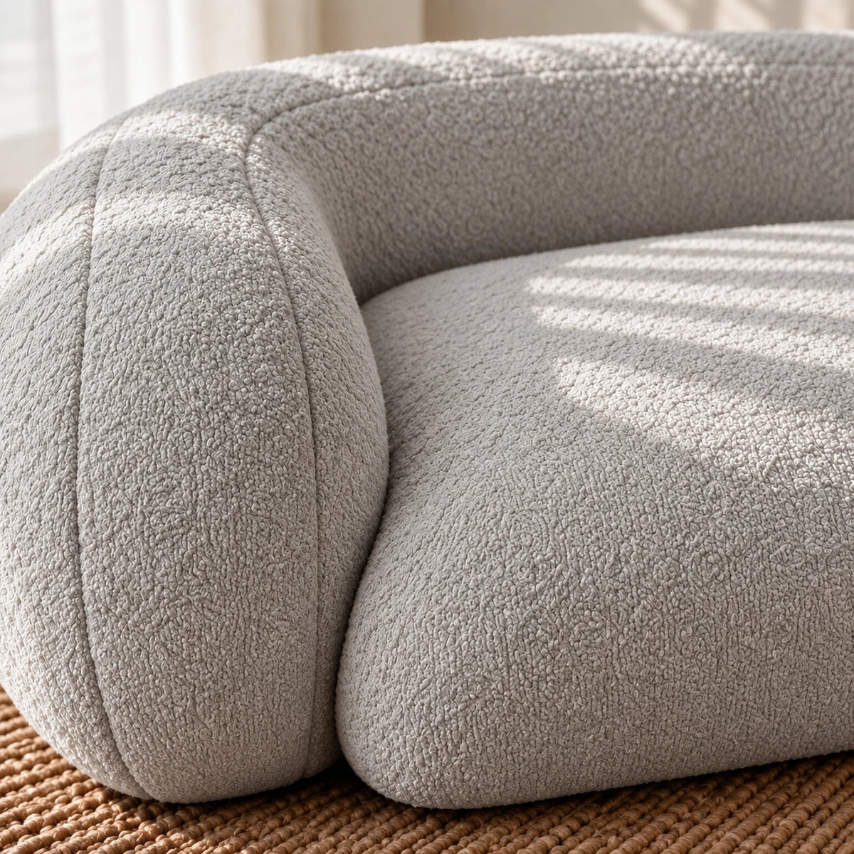 Close-up of a modern curved upholstered chair in a light warm gray bouclé fabric, resting on a woven brown rug with soft sunlight and shadow patterns across the seat.