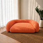 An orange bouclé pet bed with a rounded, cushioned design, placed on a brown woven rug in a sunlit room with sheer curtains and a potted plant.