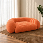 An orange bouclé pet bed with a rounded, low-profile design, placed on a woven brown rug in a sunlit room with sheer curtains and a potted plant.