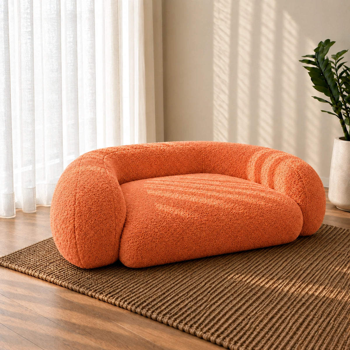 An orange bouclé pet bed with a rounded, low-profile design, placed on a woven brown rug in a sunlit room with sheer curtains and a potted plant.