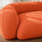 Close-up of a modern curved orange upholstered chair or sofa corner with textured boucle fabric, resting on a woven rug in soft natural light.