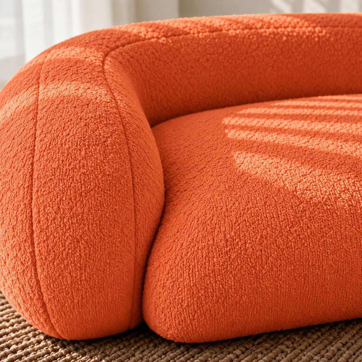 Close-up of a modern curved orange upholstered chair or sofa corner with textured boucle fabric, resting on a woven rug in soft natural light.