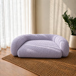 A plush, low-profile pet bed in a light lavender purple bouclé fabric, styled on a brown woven rug in a sunlit room next to a potted plant.