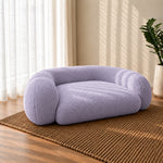 A plush, low-profile pet bed in a soft lavender bouclé fabric, styled on a brown woven jute rug in a sunlit room with sheer white curtains and a potted plant.