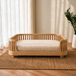 A wooden pet bed with a slatted guard and a thick textured cream cushion, placed on a woven jute rug in a sunlit room with sheer curtains and a potted plant.