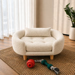 A small cream bouclé pet bed with a matching cushion and wooden legs, styled on a woven rug in a bright room with a plant and window curtains in the background.