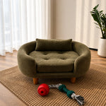 An olive green pet bed with a tufted cushion and wooden legs, styled on a woven rug in a bright room with a plant and window curtains.