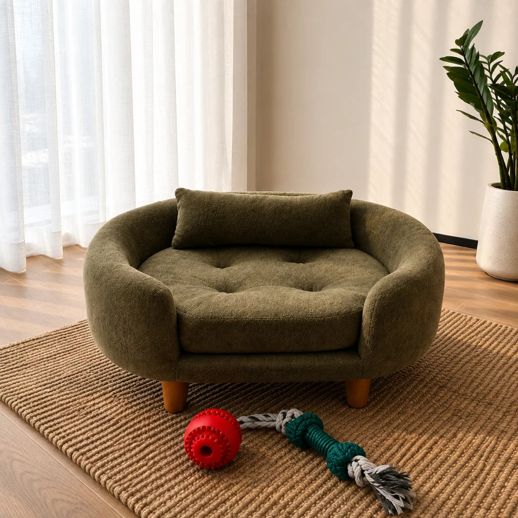 An olive green pet bed with a tufted cushion and wooden legs, styled on a woven rug in a bright room with a plant and window curtains.