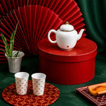White teapot and cups on a red box with a decorative fan and plant in the background