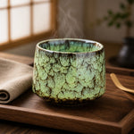 Green ceramic tea cup on a wooden tray with steam rising, set against a blurred indoor background.