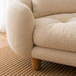 Close-up of a beige bouclé upholstered sofa corner with rounded arms and wooden legs, placed on a woven rug in a softly lit room.