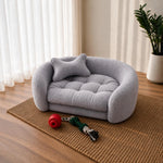 A small round gray plush pet bed with a matching pillow, placed on a woven rug in a bright room with curtains, a plant, and pet toys nearby. 