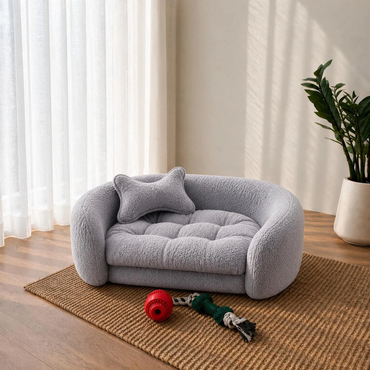 A light gray plush pet bed with a matching cushion and small pillow, styled on a woven rug in a bright room with curtains, a plant, and a red ball toy nearby. 
