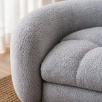 Close-up of a light gray bouclé upholstered chair or sofa corner, showing the textured fabric and tufted cushion in a softly lit interior. 
