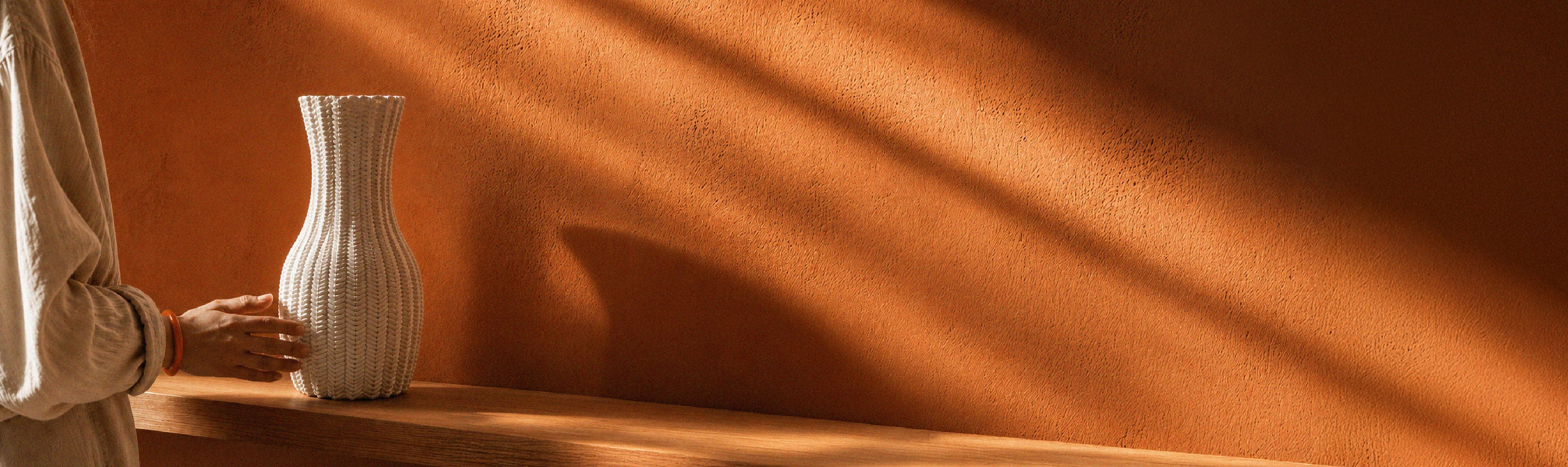White vase on a wooden shelf with warm lighting and shadow on an orange wall