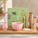Tea-making set with pink bowl, wooden spoon, and other utensils on a wooden tray.
