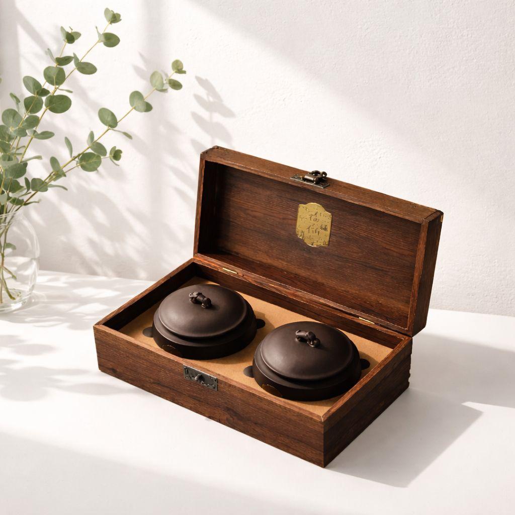 Two brown ceramic pots in a wooden box with a plant in the background