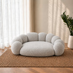 A round, low-profile pet bed with a bubbly, scalloped edge design, upholstered in a soft, textured bouclé fabric. The bed is styled in a warm, modern room setting on a woven jute rug, with sheer white curtains, warm sunlight casting shadows on the wall, and a potted plant in the background.