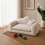 A small cream bouclé sofa with rounded arms and a matching cushion, styled in a bright living room on a woven rug with a plant and dog toy nearby.